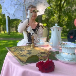 Kobieta przebrana za Blankę de Paiva. Kobieta siedzi przy małym stole. Na stole jest list, czerwona róża, porcelanowy dzbanek i filiżanki. W tle świerklaniecki park latem.