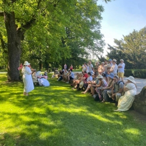Przewodniczka stojąca przy kobiecie przebranej za Blankę de Paiva. Publiczność skierowana w stronę przewodniczki. W tle świerklaniecki park latem.