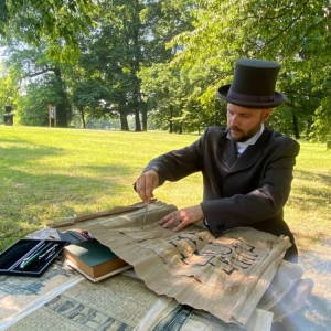 Mężczyzna przebrany za architekta siedzi przy stole. Na stole są rozrzucone papiery i dokumenty. W tle świerklaniecki park latem.